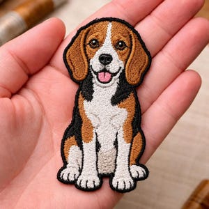 May include: An embroidered iron-on patch featuring a beagle dog in a sitting position. The dog has brown, black, and white fur, dark brown eyes, a black nose, and an open mouth with a pink tongue visible. The patch has a black stitched border.
