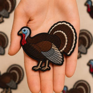 May include: An embroidered iron-on patch shaped like a turkey. The turkey features a dark brown body, a light brown and cream fan-shaped tail, a blue head, a red wattle, and tan legs. The entire design is outlined in a thick black border.