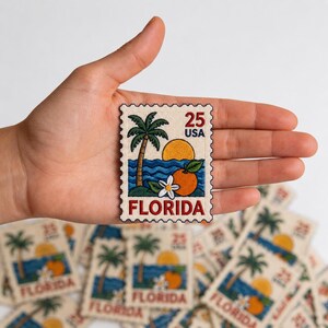 May include: A rectangular embroidered patch designed like a postage stamp featuring a palm tree, a yellow sun, blue ocean waves, an orange, and a white blossom. The text 25 USA and FLORIDA is stitched in red and black on a cream-colored fabric background.