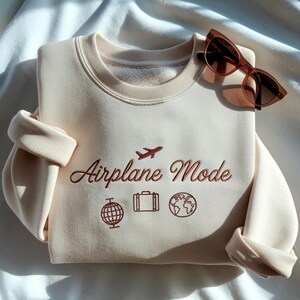 Może przedstawiać: Kremowa bluza z haftem "Airplane Mode" i ikonami podr&oacute;żniczymi: samolot, walizka, globus. Na wierzchu leżą brązowe okulary przeciwsłoneczne. Złożone rękawy i miękki materiał sugerują swobodny, casualowy styl.