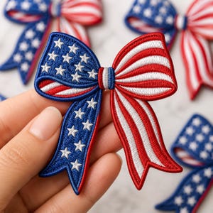 May include: Embroidered bow with a patriotic design featuring red and white stripes and blue sections with white stars. The bow is held in a hand, with other bows visible in the background. The bows are likely for crafts or decorations.