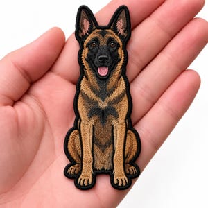 May include: An embroidered patch featuring a German Shepherd dog in a sitting position. The dog has a black muzzle, tan and brown fur, pointed ears, and an open mouth with a pink tongue visible. The patch has a black border and detailed stitching.