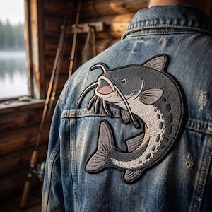 May include: A blue denim jacket featuring a large, embroidered patch on the back. The patch depicts a stylized dark gray and white catfish with long whiskers, a pinkish open mouth, and a curved body shape, stitched directly onto the fabric.