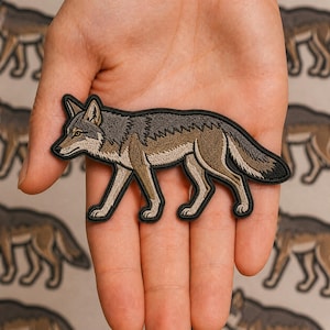 May include: An embroidered patch featuring a side profile of a wolf in shades of gray, brown, and cream. The wolf is depicted in a walking stance with a black border outlining its entire shape, showcasing detailed stitching throughout the design.