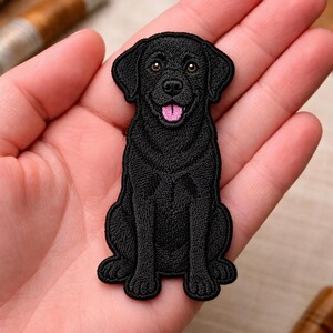 May include: An embroidered patch featuring a black Labrador retriever dog sitting in a forward-facing position. The dog has brown eyes, a black nose, and a pink tongue visible in its open mouth. The embroidery uses dense black thread.