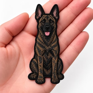 May include: An embroidered patch featuring a brindle Dutch Shepherd dog sitting in an upright position. The dog has a black muzzle, pointed ears, and a pink tongue visible. The patch has a black border and detailed brown and black fur texture.
