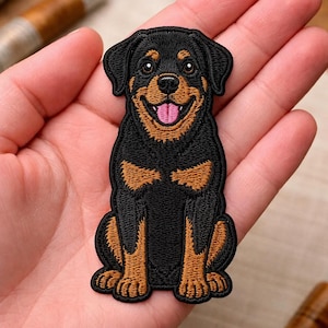 May include: An embroidered patch featuring a Rottweiler dog sitting in a front-facing position. The dog has black fur with tan markings on its face, chest, and paws, a black nose, and an open mouth showing a pink tongue and white teeth.