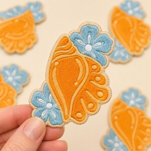 May include: Embroidered patches in a butterfly wing design. The patches feature a vibrant orange wing shape with light blue floral accents and a cream-colored border. The patches are suitable for clothing or craft projects.