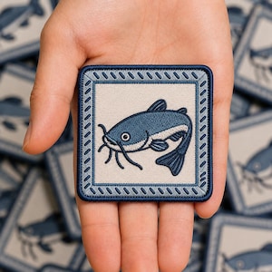 May include: A square embroidered patch featuring a blue catfish illustration centered on a light beige background. The patch is framed by a dark blue border with a diagonal stitched pattern, creating a distinct square shape with rounded corners.