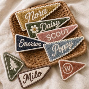 May include: A collection of triangular pennants with textured surfaces, featuring embroidered names including Nora, Daisy, Scout, Emerson, Poppy, and Milo, alongside a pine tree icon and a letter W, all arranged on a woven square tray.