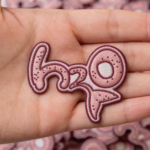 May include: Embroidered patch in pink and burgundy with the text "H2O". The patch has a cursive font and a decorative border. The patch is held in a hand.