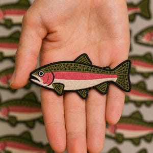 May include: An embroidered iron-on patch shaped like a rainbow trout fish. The fish features a dark olive green back with black spots, a vibrant pink horizontal stripe along the side, and a cream-colored belly, all outlined in a thick black border.