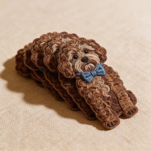May include: A stack of brown dog-shaped coasters with a blue bow tie. The coasters are made of a textured material, with detailed stitching to create the dog's fur. The dog's face has a black nose and eyes.