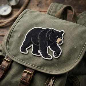 May include: An olive green canvas backpack featuring a black bear embroidered patch on the front flap. The backpack includes brown leather straps with brass-toned metal buckles and a top handle, showcasing a textured fabric construction.