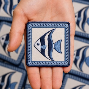 May include: A square embroidered patch featuring a stylized angelfish in shades of navy blue, light blue, and white. The fish is centered on a cream background, enclosed by a decorative navy blue border with a repeating diagonal stitch pattern.