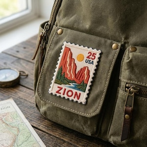 May include: An olive green canvas backpack featuring a rectangular embroidered patch shaped like a postage stamp. The patch depicts a landscape with red rock formations, a yellow sun, and a blue river, with the text 25 USA and ZION.