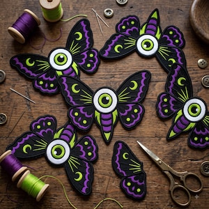 May include: Five embroidered moth patches featuring a central eye design with black, purple, and lime green thread. The patches are arranged with sewing supplies including purple and green thread spools, metal needles, buttons, and scissors.