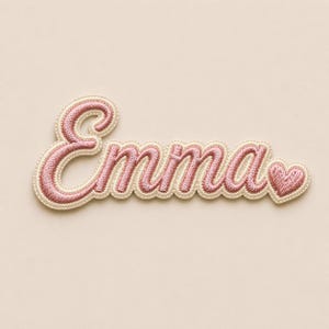 May include: Embroidered patch in script font, spelling "Emma" in pink thread. The name is outlined in a cream-colored border with a scalloped edge. A small pink heart is at the end of the name.