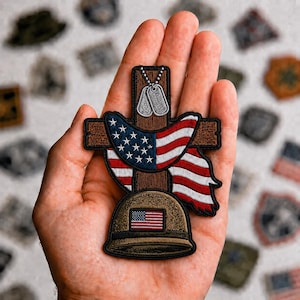May include: A fabric patch featuring a brown wooden cross draped with an American flag, a tan military helmet with a small American flag emblem on the front, and a pair of silver-toned metal dog tags hanging from the top of the cross.