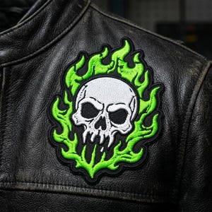 May include: An embroidered patch featuring a white skull with black outlines surrounded by bright green flames, stitched onto a black leather jacket. The skull design has deep black eye sockets and a detailed jawline, framed by a black border.