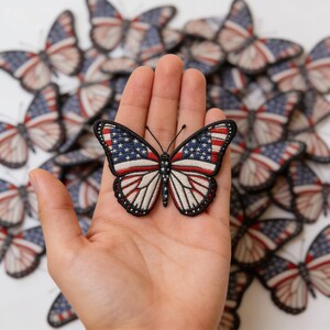 May include: Embroidered butterfly patch featuring the American flag design. The butterfly has blue wings with white stars, red and white stripes, and a black border. A collection of similar patches is in the background.