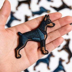 May include: Embroidered patch featuring a Doberman Pinscher dog in shades of black, blue, and brown. The patch has a detailed design with a black outline and is likely for application on clothing or accessories.
