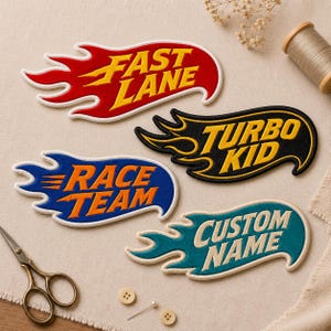 May include: Four embroidered flame-shaped patches with white borders. The red patch reads FAST LANE in yellow, the black patch reads TURBO KID in yellow, the blue patch reads RACE TEAM in orange, and the teal patch reads CUSTOM NAME in white.