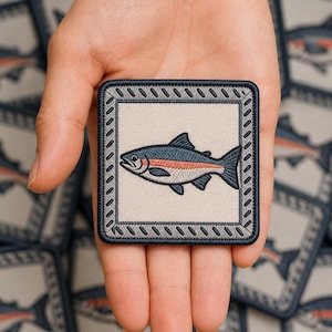May include: A square embroidered patch featuring a central illustration of a fish with a dark blue back, a light underbelly, and a horizontal reddish-orange stripe along its side, enclosed by a dark blue border with a diagonal stitched pattern.
