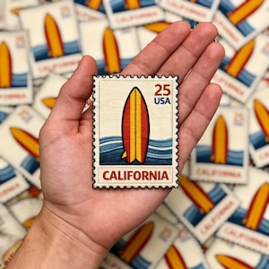 May include: A rectangular stamp featuring a surfboard design with red, yellow, and orange stripes. The stamp reads "25 USA" and "CALIFORNIA" in bold letters. The background shows a blue ocean with white waves.