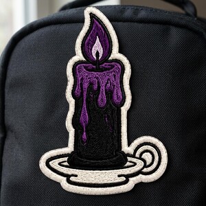 May include: An embroidered patch featuring a black candle in a white candle holder. The candle has purple wax dripping down its sides and a purple flame at the top. The patch has a thick white border and is attached to a black fabric surface.