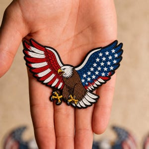 May include: An embroidered patch featuring a bald eagle with wings spread wide. The left wing displays red and white stripes, while the right wing features a blue field with white stars. The eagle has a brown body, white head, and yellow talons.
