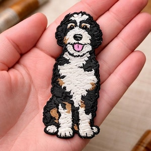 May include: Embroidered patch featuring a Bernedoodle dog. The patch has a black, white, and brown color scheme, with a smiling face and pink tongue. The dog is sitting upright, with its paws visible.