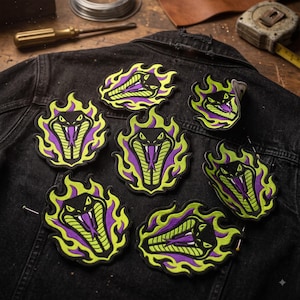 May include: Seven embroidered patches featuring a black cobra head with a purple tongue, surrounded by bright lime green flames. The patches are arranged on a black denim jacket. Each patch displays a stylized snake with sharp fangs.