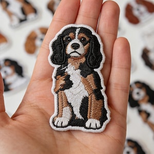 May include: Embroidered patch featuring a Cavalier King Charles Spaniel dog. The patch has a white border and depicts the dog in black, brown, and white. The dog is sitting with its front paws forward. Other dog patches are visible in the background.