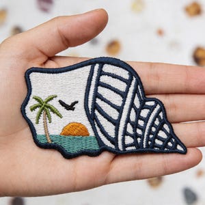 May include: Embroidered patch featuring a seashell design with a coastal scene. The patch includes a palm tree, a setting sun, and a flying bird, all outlined in navy blue. The background is white and teal.