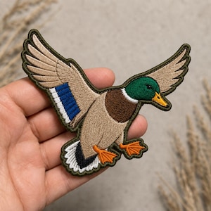 May include: An embroidered patch featuring a mallard duck in flight with wings spread wide. The duck has a dark green head, a brown chest, tan wings with a bright blue and white speculum, and orange webbed feet against a dark green border.