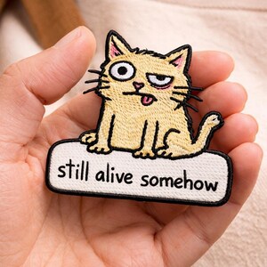 May include: Embroidered patch featuring a cartoon cat with a slightly annoyed expression, tongue sticking out. The patch is light yellow with black details and the text "still alive somehow" in black on a white background.