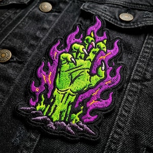 May include: An embroidered patch featuring a bright green monster hand with sharp white claws emerging from the ground, surrounded by vibrant purple flames with yellow highlights, all stitched onto a black denim fabric background.