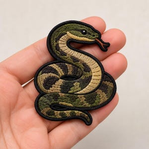 May include: An embroidered patch featuring a coiled snake with a black border. The snake has a green and brown patterned body, a light tan underbelly, and a red forked tongue. The design is detailed with dark stitching throughout the scales.