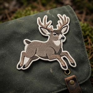 May include: An embroidered patch featuring a brown deer with large antlers in a leaping pose. The deer has white accents on its face, neck, and underbelly, with black hooves and a white border. The patch is attached to a dark olive green fabric.