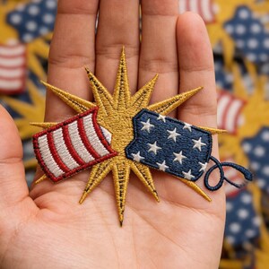 May include: Embroidered patch featuring a gold starburst design with two flag-like elements. One element has red and white stripes, the other has white stars on a blue background. The patch is held in a hand.