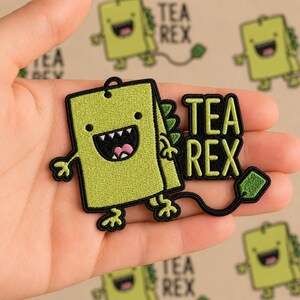 May include: Embroidered patch featuring a green cartoon dinosaur with the text "TEA REX". The dinosaur has a smiling face, small arms and legs, and a tea bag tail. The patch is on a tan background.