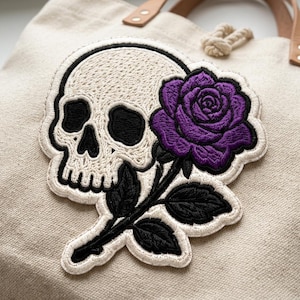 May include: A beige canvas tote bag featuring an embroidered patch of a white skull with black eye sockets and nasal cavity, paired with a vibrant purple rose on a black stem with black leaves, all outlined in a thick white border.