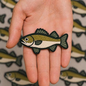 May include: An embroidered iron-on patch shaped like a fish with olive green scales on the top half, a cream-colored belly, and dark green fins and tail. The fish has a black outline, a white eye with a black pupil, and a small open mouth.