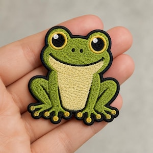 May include: An embroidered patch featuring a green frog with a light yellow belly, large black eyes with yellow outlines, and small white highlights. The frog has a black border and is depicted in a seated position with visible toes and fingers.