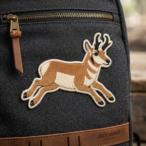 May include: A dark charcoal gray canvas backpack featuring an embroidered patch of a tan and white pronghorn antelope in a running pose. The bag includes a brass-colored metal zipper with a brown leather pull tab and a brown leather base label.