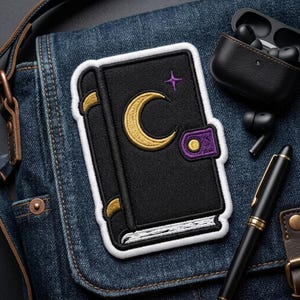 May include: An embroidered patch featuring a black book with a gold crescent moon and a small purple star on the cover. The book has a purple clasp with a gold dot, white page edges, and gold accents on the spine, all outlined in a thick white border.