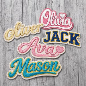 May include: Embroidered name patches in various colors and fonts, including "Olivia," "Oliver," "Ava," "Jack," and "Mason." Each patch features a unique design with a cursive font and a decorative border. The patches are displayed on a wooden surface.