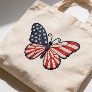 May include: A cream-colored tote bag featuring an embroidered butterfly design. The butterfly's left wing is blue with white stars, and the right wing is red and white striped, resembling the American flag. The bag has two handles.