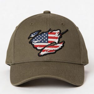 May include: Olive green baseball cap featuring an embroidered American flag design. The flag appears torn, with the stars and stripes visible. The cap has a curved brim and a structured crown.